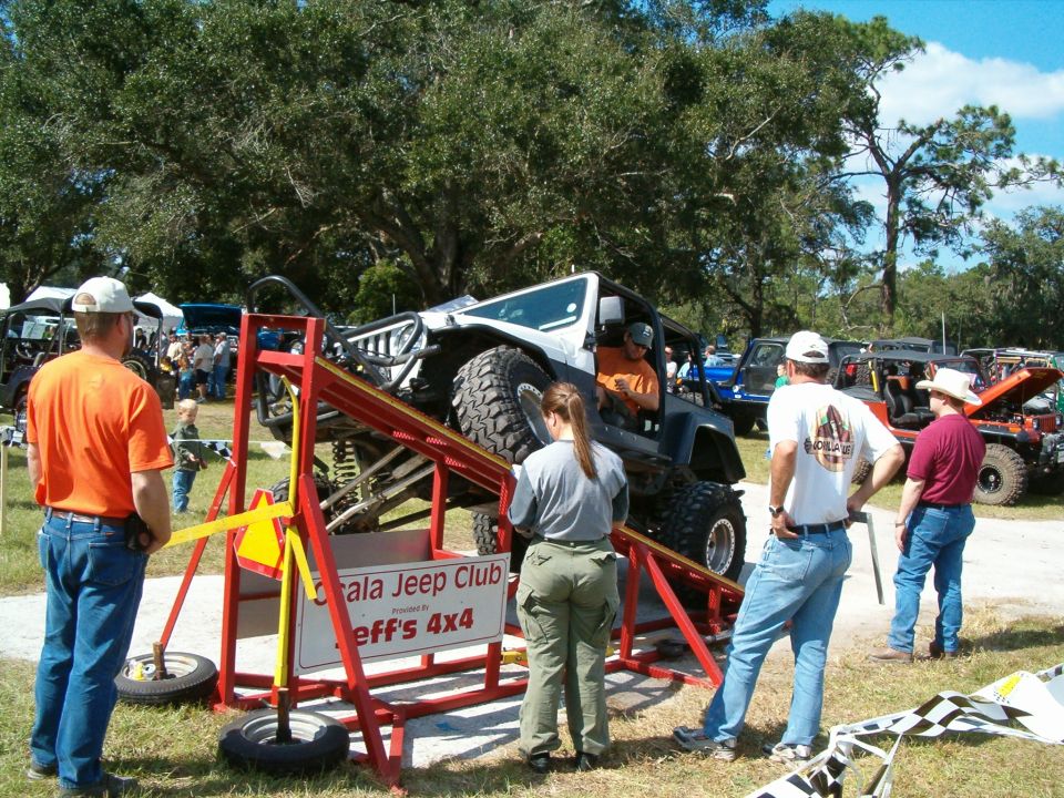 Modified Jeep Wrangler on a red ramp, with people watching. The vehicle has large tires and a lifted suspension