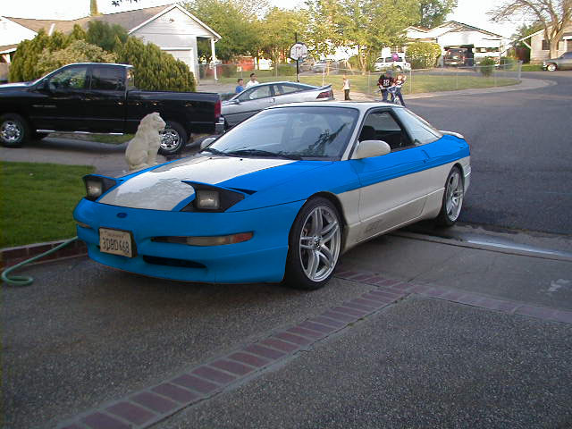 Blue and white 1997 Ford Probe parked in driveway. Black pickup truck and a dog are nearby