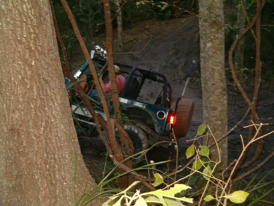 Green SUV with top off tilted on a muddy wooded trail, seen through tree trunks, brake light lit