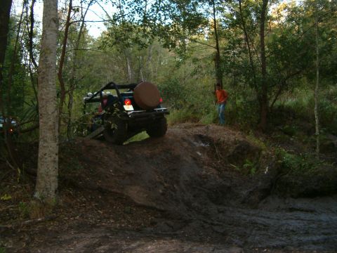 1991 Ford Bronco - An off-road vehicle navigates a challenging muddy incline surrounded by dense woodland.
