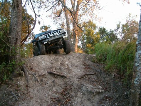 1991 Ford Bronco - A Jeep Cherokee XJ navigates a steep, muddy incline with tall grass and trees in the background.