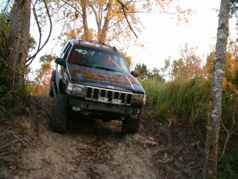 1991 Ford Bronco - A black Jeep Grand Cherokee navigates a steep dirt trail surrounded by trees and tall grass.