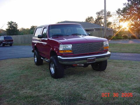 1989 Ford Bronco - Front three-quarter view of a lifted dark red full-size Ford Bronco with a white hardtop, parked on a grass lawn at sunset next to a drivew…