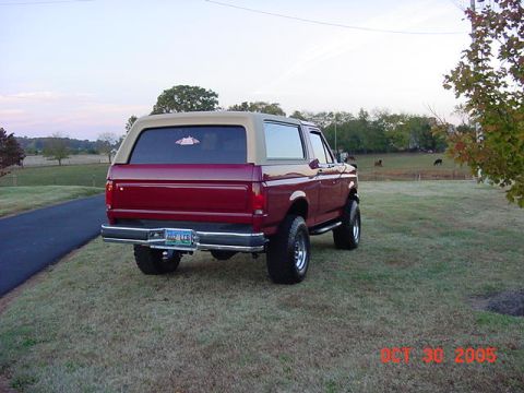 1989 Ford Bronco - Rear three-quarter view of a red two-door Ford Bronco with a tan removable hardtop, parked on grass beside a driveway with pasture, cattle…