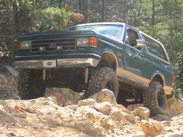 Green Ford Bronco with Ford grille emblem climbing boulders on a rocky wooded trail