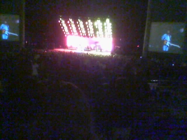 Concert stage with bright lights and two large screens showing performers, viewed from a dark audience area
