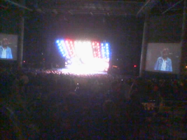 Concert stage with large screens showing performers, crowd in foreground
