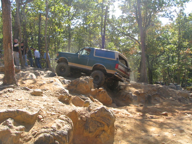 Blue and tan lifted Ford Bronco crawling over rocks on a wooded trail, spectators watching