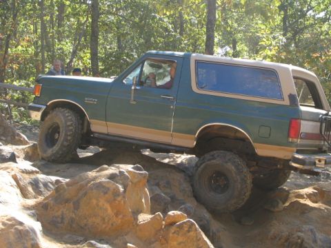1990 Ford Bronco - This 1990 Ford Bronco is shown tackling a challenging off-road course with large rocks and trees in the background.