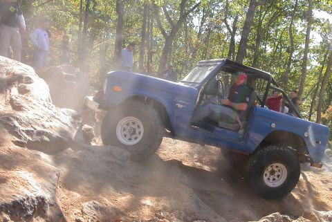 1990 Ford Bronco - A blue Ford Bronco navigates a challenging rocky terrain with spectators observing the off-road adventure.