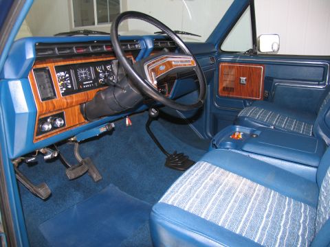 1984 Ford Bronco - The interior of a vintage Ford truck shows a blue dashboard with wood grain accents and patterned cloth seats.