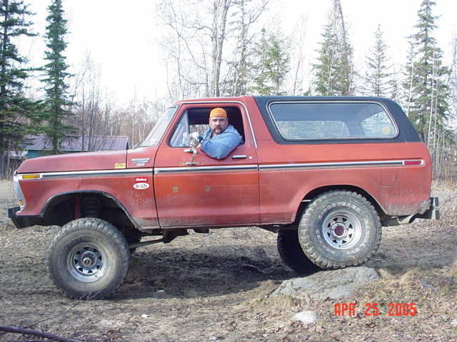 Lifted red Ford Bronco in profile on dirt, driver leaning out the window, spruce trees behind