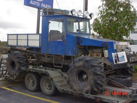 1976 Jeep CJ7 - This custom-built off-road vehicle, with its distinctive large tires and blue cab, is seen loaded onto a trailer. A sign for "AUTO PARTS" i…