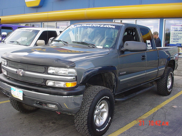 Dark gray pickup truck with a black bug deflector and a banner on the windshield that says TRAILTOYZ.COM. Another white truck is parked next to it