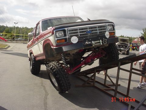 1976 Jeep CJ7 - A lifted Ford Bronco is positioned on metal ramps, with other vehicles visible in the background.
