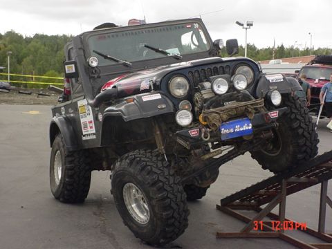 1976 Jeep CJ7 - Modified black Jeep CJ with aggressive off-road tires is positioned on a ramp, with a "Team Alaska" sticker on the front.