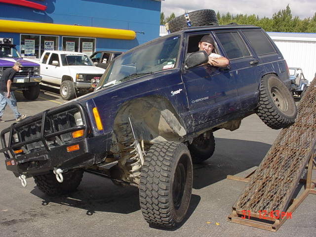 Blue Jeep Cherokee XJ with off-road tires on a ramp, front bull bar, spare tire on roof