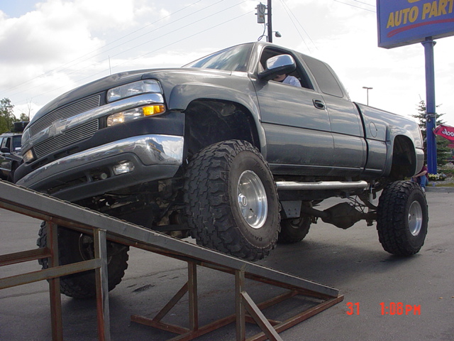 Lifted dark gray pickup truck with large tires on metal ramps, driver visible