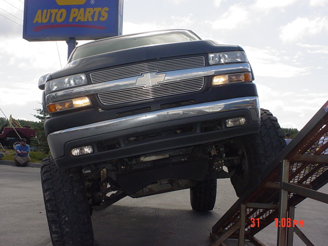 Dark Chevrolet Silverado truck with chrome grille, front end tilted up on a ramp. Auto Parts sign in background