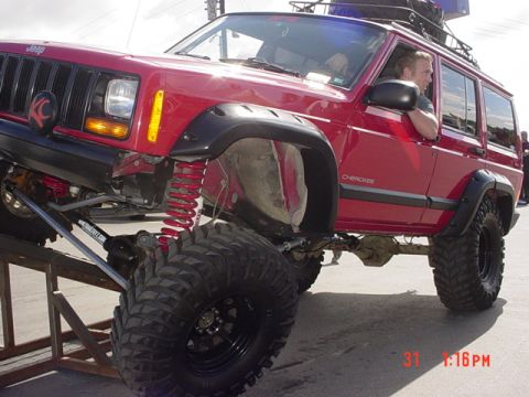 1976 Jeep CJ7 - A modified red Jeep Cherokee SUV is shown with its front wheel elevated on a ramp, highlighting its off-road capabilities.
