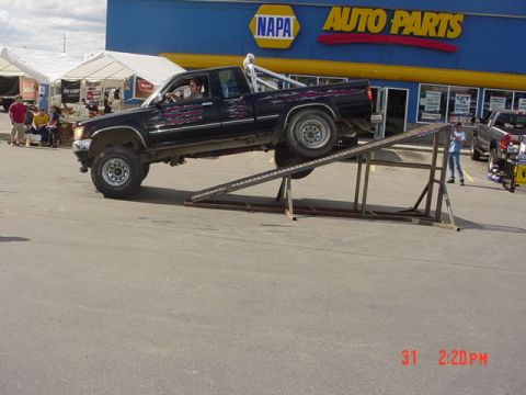 1976 Jeep CJ7 - A black pickup truck is positioned on a ramp outside a NAPA Auto Parts store, with other vehicles and tents visible in the background.