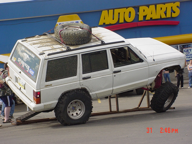 White Jeep Cherokee SUV with large tires, lifted suspension, and a spare tire on the roof rack, on display in front of an Auto Parts store
