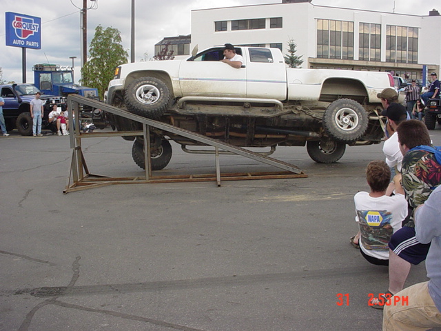 White lifted pickup flexing on a steel ramp at a parking lot event with spectators