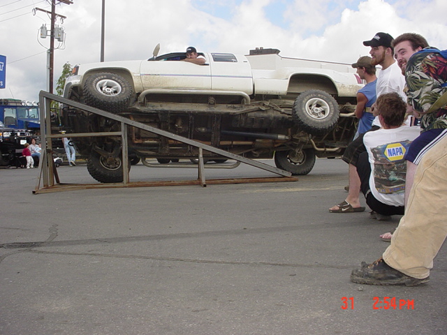 White lifted pickup climbing a steel ramp obstacle as spectators watch