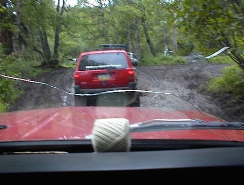 1978 Ford Bronco - A red SUV navigates a muddy off-road path through a wooded area, with another vehicle visible ahead.