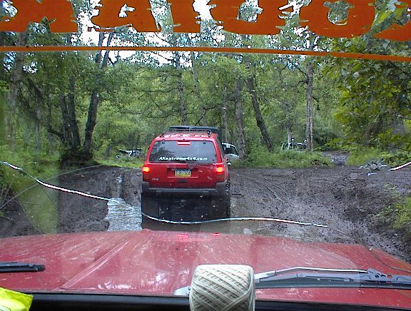 View from inside a vehicle looking at a red SUV on a muddy forest road. Another vehicle is visible in the distance
