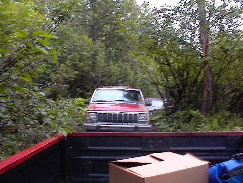 1978 Ford Bronco - A red Jeep Cherokee is seen on a wooded road, with another vehicle partially visible behind it.