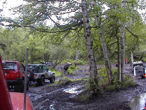 Several off-road vehicles navigate a muddy, wooded trail