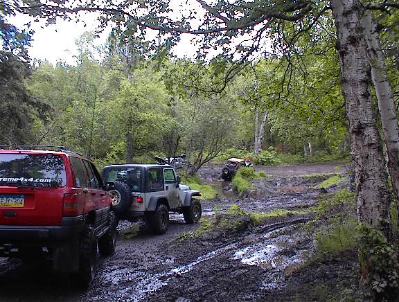Red SUV and silver Jeep on muddy track with trees and other vehicles in background