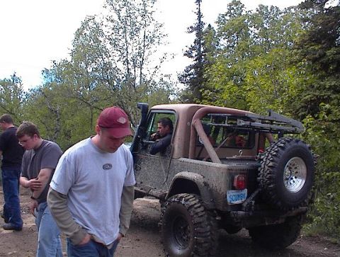1978 Ford Bronco - A group of people stand around a dirty off-road vehicle parked on a dirt path surrounded by trees.