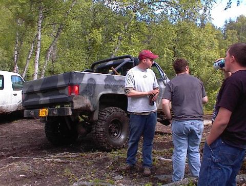 1978 Ford Bronco - A camouflaged pickup truck with large off-road tires is parked on a dirt path, with several people standing nearby.