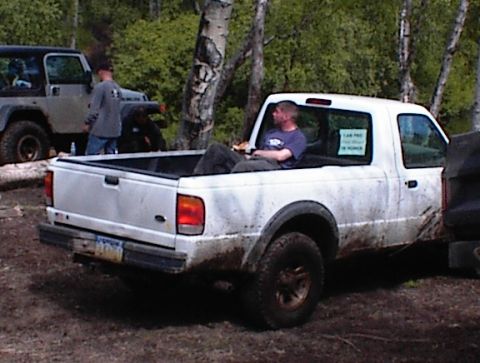 1978 Ford Bronco - A white pickup truck and a Jeep are parked off-road, with a man relaxing in the truck bed.
