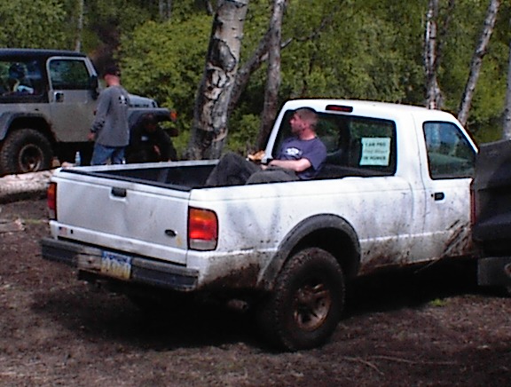 White pickup truck with a man sitting in the bed, parked next to a Jeep. Mud on the truck's body and wheels