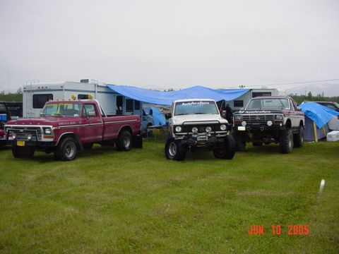 1978 Ford Bronco - Three lifted trucks and RVs are parked on a grassy field under a cloudy sky.