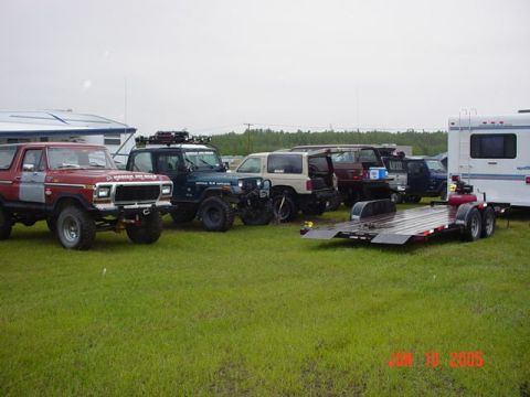 1978 Ford Bronco - Several off-road vehicles and a trailer are parked on a grassy field under an overcast sky.