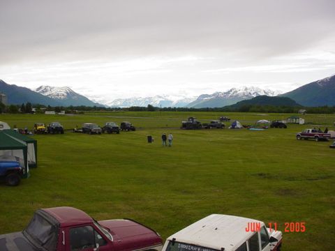 1978 Ford Bronco - A gathering of off-road vehicles and tents is set up in a grassy field, with snow-capped mountains visible in the distance under an overcas…