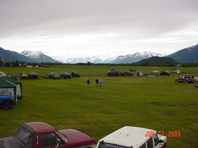 Field gathering with multiple SUVs, trucks, tents, and mountains under a cloudy sky