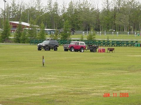 1978 Ford Bronco - Two SUVs and a trailer are parked on a grassy field with trees and picnic tables in the background.