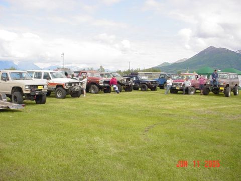 1978 Ford Bronco - A group of off-road vehicles are gathered in a field on a cloudy day, with mountains visible in the distance.