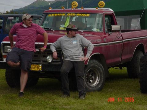 1978 Ford Bronco - A red pickup truck is parked on grass with two men posing next to it. The truck has a custom decal and lights on the roof.