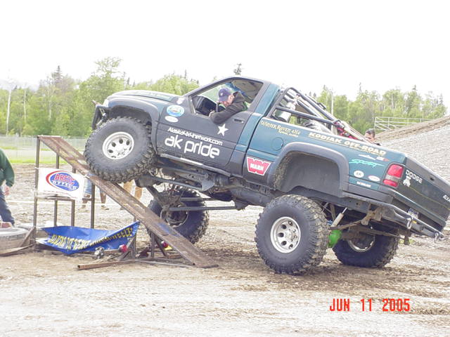 Modified Dodge pickup truck with large tires climbing a ramp on an obstacle course. Text on the truck says 'ak pride' and 'KODIAK OFF-ROAD'