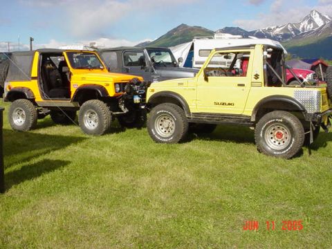 1978 Ford Bronco - Two Suzuki Samurai SUVs are parked on a grassy field with mountains visible in the distance.
