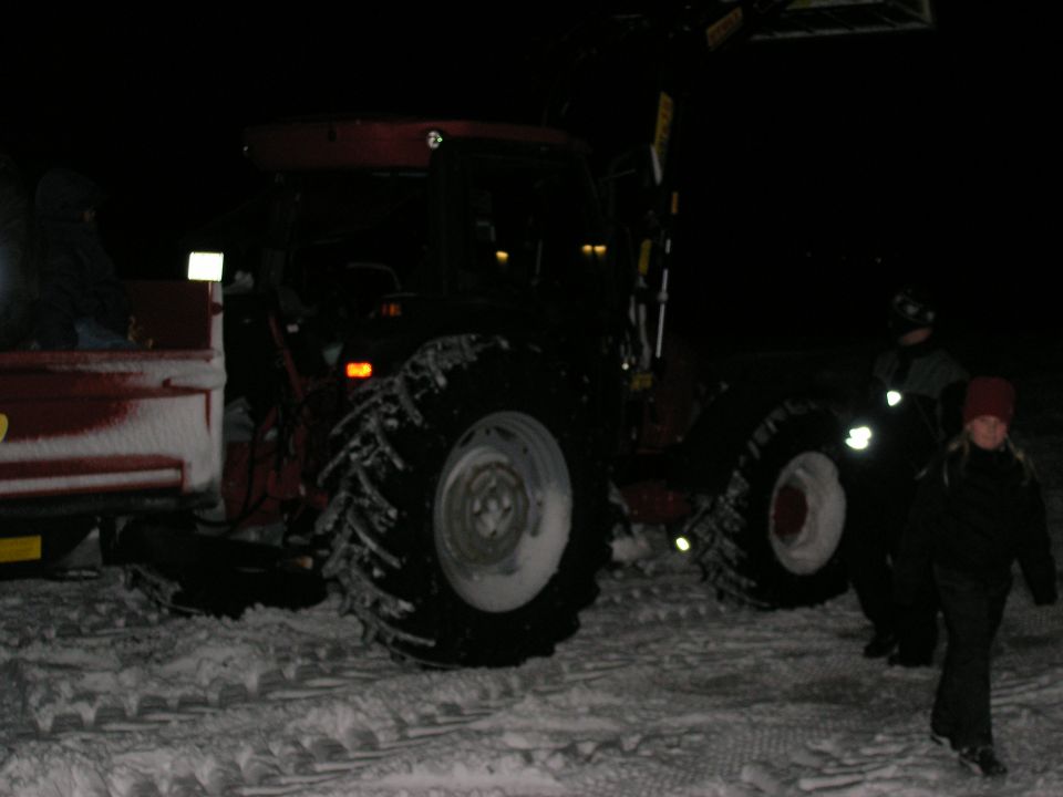 Tractor with large tires in snow at night, towing a red trailer. People are visible