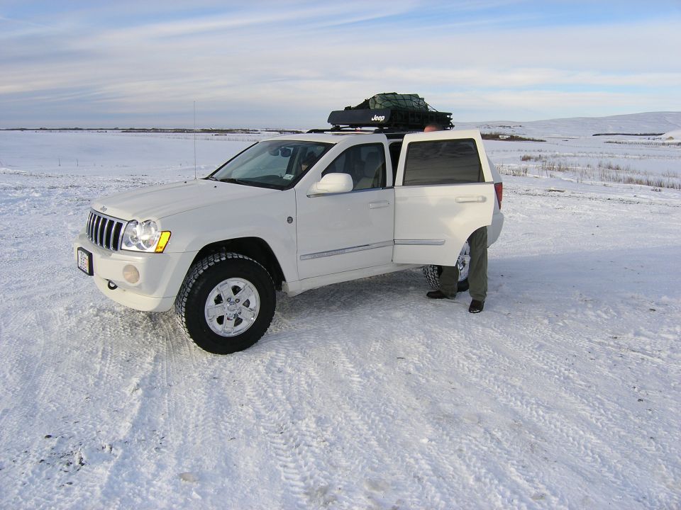 White Jeep Grand Cherokee, 2005 model, with open doors and roof rack loaded with gear, parked in a snowy field