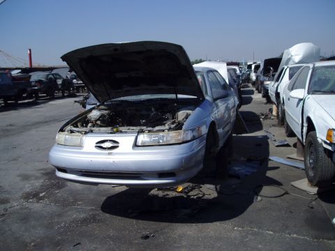 1993 Ford Taurus - Junkyard scene with a silver sedan, front end visible, hood up, and other vehicles in the background.