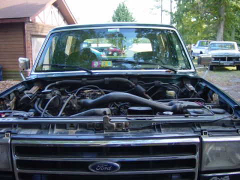 1991 Ford Bronco - The engine bay of a dark green 1991 Ford Bronco is shown with the hood open. Other vehicles are parked in the background.